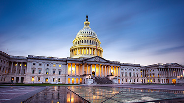 The US Capitol building at dusk, with dramatic lighting highlighting its iconic dome and neoclassical architecture against a clear blue sky.