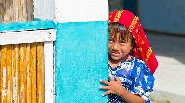 A child with a headdress on peeking out from behind a door and smiling.