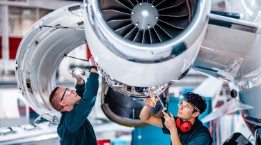 Two maintenance engineers inspect a jet engine inside an aircraft maintenance hangar.