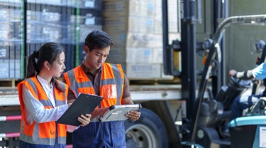 Warehouse workers double-check paperwork as forklift loads shipment into a truck.