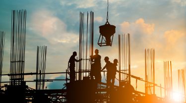 Four construction workers silhouetted against a sunset sky, actively working on a construction site.