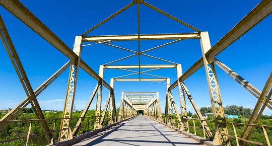 A yellow steel bridge stretches towards a heavily treed area.