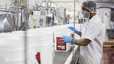 A man dressed in white moves containers of feta cheese on a conveyer belt.