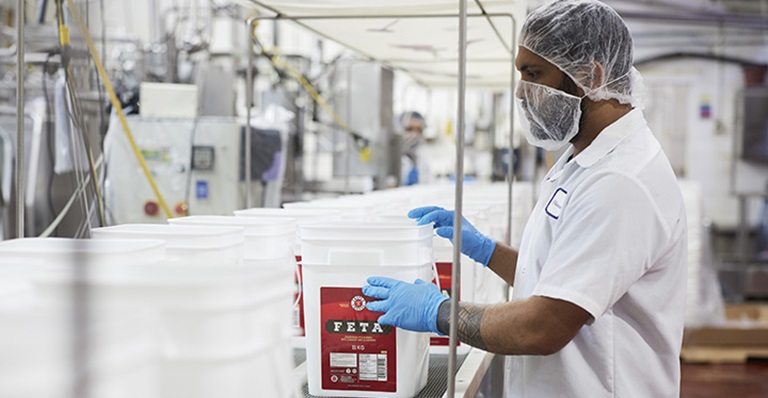 A man dressed in white moves containers of feta cheese on a conveyer belt.