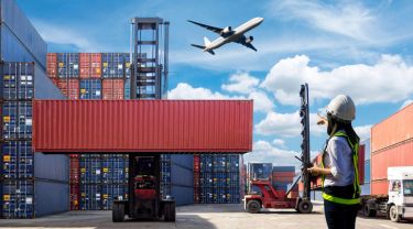 An airplane flying above shipping containers, trucks, and a worker wearing a safety vest on the ground.