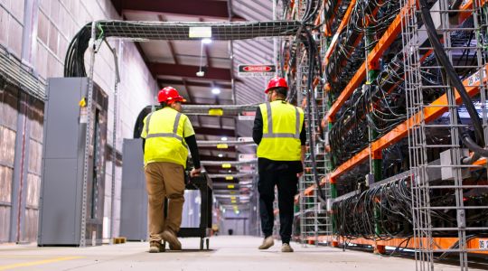 Deux personnes portant des casques de sécurité et des gilets haute visibilité se déplacent dans une allée de centre de données industriel bordée de baies de serveurs et de câbles.