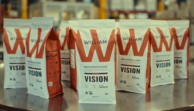 Packaged Café William espresso beans in focus with a factory background softly blurred.