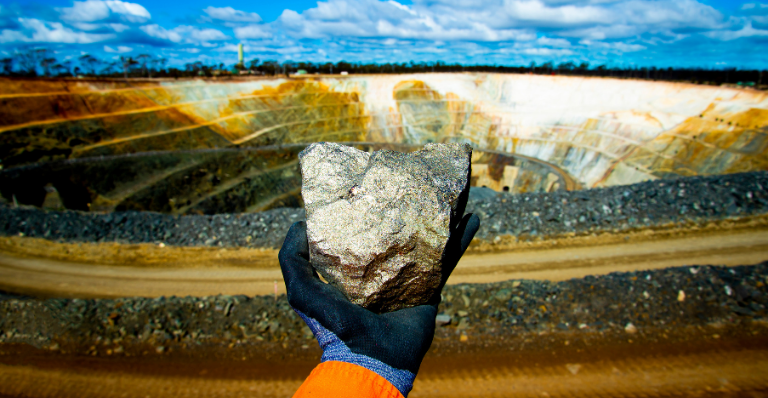 Main gantée tenant un gros minerai de nickel sulfuré devant une fosse minière à ciel ouvert.