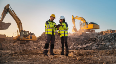 Female and male civil engineers review a tablet at a mine site, with two excavators operating in the background.