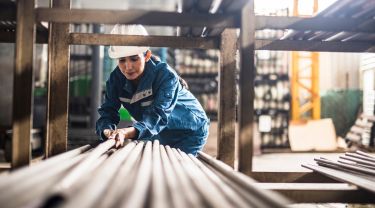 A woman wearing coveralls and a safety helmet bending down to move steel poles from a shelf.