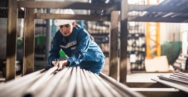 A woman wearing coveralls and a safety helmet bending down to move steel poles from a shelf.