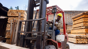 Forklift driver in yellow hard hat operates red forklift in lumber yard amid stacks of wooden boards.
