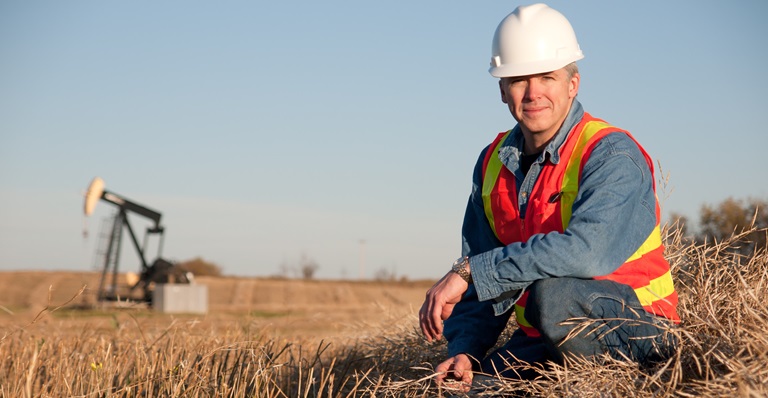 Albertan engineer in safety gear inspects soil near oil well pumpjack