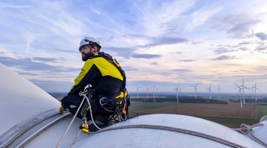 German wind turbine technician secured on nacelle overlooking a vast wind farm under a cloudy sky