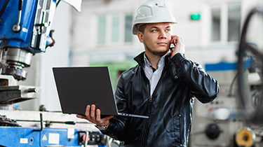 An engineer standing in front of an industrial machine with his laptop talks on the phone