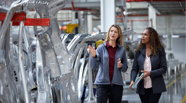 Two female engineers chat near car chassis in factory.