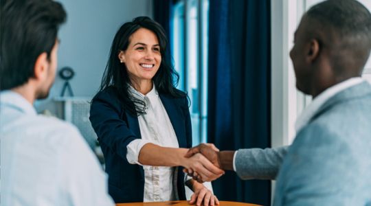 A smiling business professional shaking hands with another, while a third person stands beside them facing away from the camera.