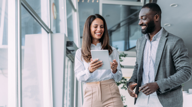 Two black professionals, smiling and collaborating over a tablet while walking in a modern office.