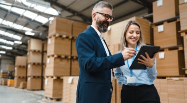 A businessman signs a document on a tablet held by a businesswoman in a warehouse.