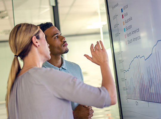 Two business people standing in front of a presentation screen discussing quarterly revenue. 