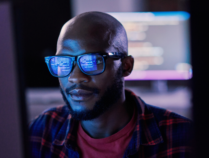 A man sitting in a dark room in front of his computer, lines of code reflecting off his glasses.