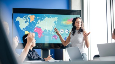 Canadian businesswoman stands before a data display showing a colourful world map with arrows pointing across the globe.