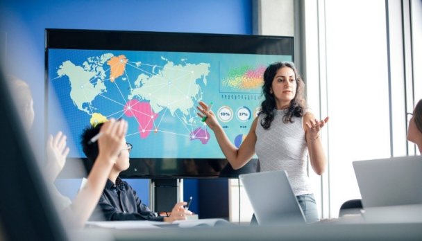 Canadian businesswoman stands before a data display showing a colourful world map with arrows pointing across the globe.