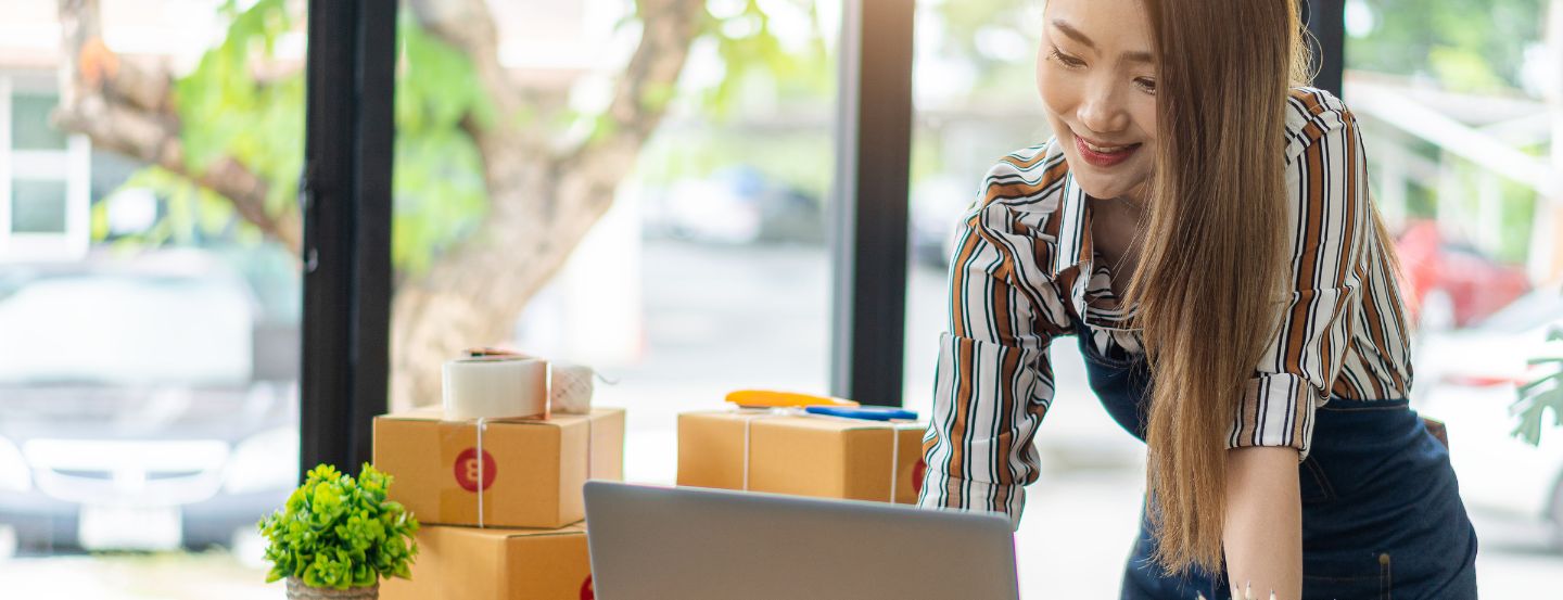 Une femme d'affaires debout devant un bureau, utilisant un ordinateur portable, avec de petites boîtes à proximité.
