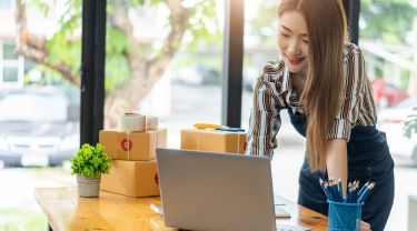 Une femme d'affaires debout devant un bureau, utilisant un ordinateur portable, avec de petites boîtes à proximité.