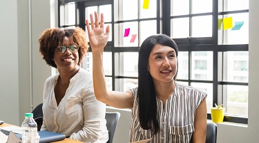 Two women engaged in meeting – one with raised hand.  