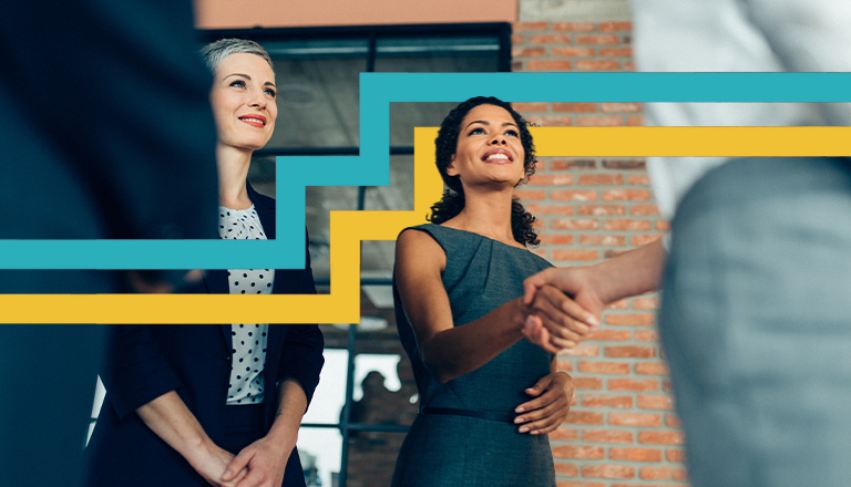 Two women smiling and facing two other people. One of the women is shaking hands with one person.