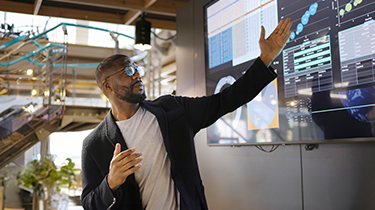 Stock image of a Black man conducting a seminar / lecture with the aid of a large screen. The screen is displaying graphs & data associated with movie clips of the earth.