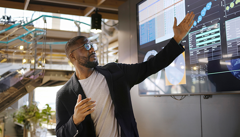 Stock image of a Black man conducting a seminar / lecture with the aid of a large screen. The screen is displaying graphs & data associated with movie clips of the earth.