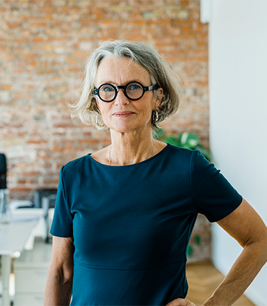 Une femme élégamment vêtue, avec des cheveux gris et des lunettes, se tient avec assurance dans un bureau.