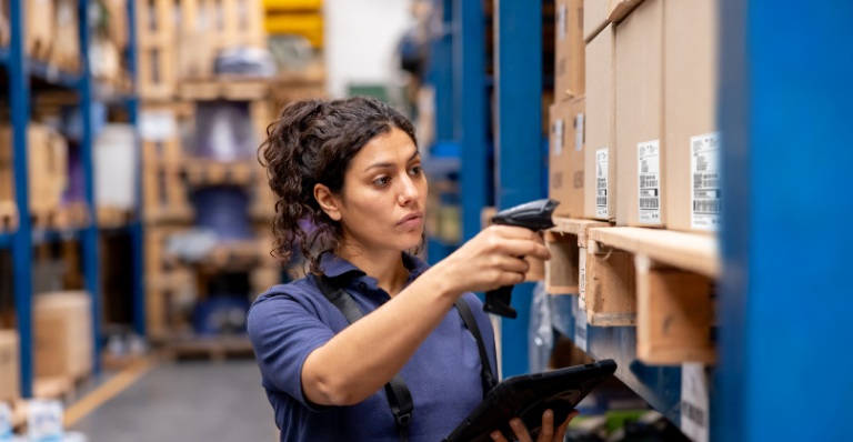 Worker scans inventory with barcode scanner and tablet in a warehouse aisle.