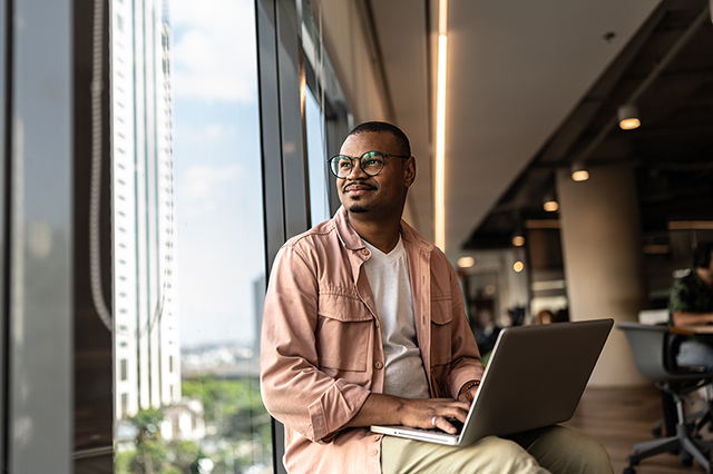 A Black businessman contemplating in the office, looking out the window with a silver laptop on his lap.