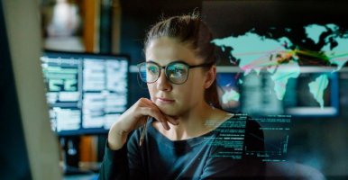 Une jeune femme portant des lunettes assise dans un bureau sombre et entourée d’écrans d’ordinateur. En face d’elle se trouve un écran transparent montrant une carte du monde avec des données en superposition.