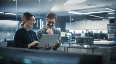 Two colleagues collaborating over a laptop inside a technology workspace filled with computer screens.
