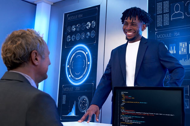 A professional discusses data with a customer using large digital dashboards in a modern office.