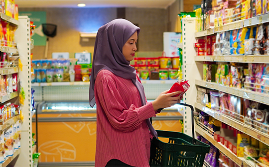 A young Muslim woman in a grocery aisle, carefully reading the label on a food item to determine if it is halal certified. The halal food industry is an opportunity for Canadian companies to diversify their product offering