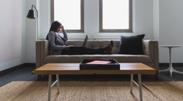 Person sitting on a sofa with a laptop, working in a bright, minimalist living room with large windows, a coffee table, and neutral decor.