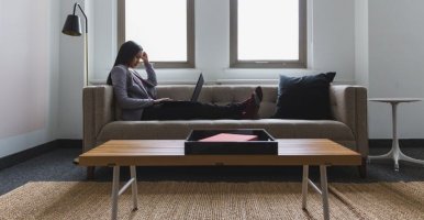 Person sitting on a sofa with a laptop, working in a bright, minimalist living room with large windows, a coffee table, and neutral decor.