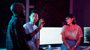 Trois personnes échangent autour d’écrans d’ordinateur dans un espace de travail technologique éclairé en lumière tamisée.