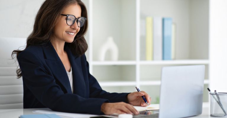 Une femme d'affaires souriante portant des lunettes travaillant sur un ordinateur portable à un bureau dans un bureau moderne.