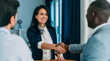 A businesswoman and a businessman shaking hands in an office, with another businessman standing nearby, observing the interaction. 