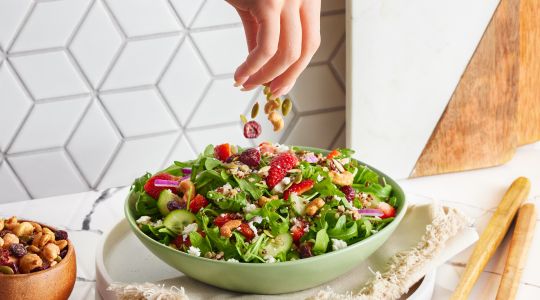 A woman’s hand adding salad toppings to a bowl of salad, with a bowl of mixed nuts on the side in a modern kitchen.