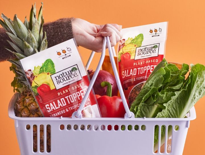 A hand holding a grocery basket filled with fresh produce and plant-based food packages