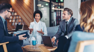 Quatre personnes assises dans un bureau de style lounge en train de discuter autour d'une petite table avec une tasse, un ordinateur portable et un carnet.