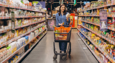 Une femme debout dans l'allée d'une épicerie, tenant un produit et poussant un chariot.