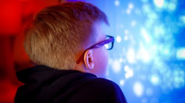 A young boy wearing glasses looks at a glowing screen in a red LED-lit space.